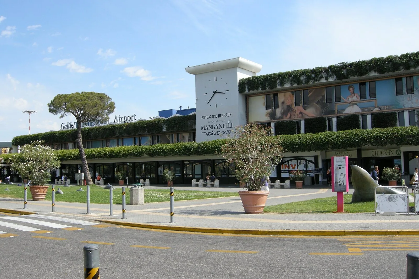 Facciata del terminal dell'aeroporto Galileo Galilei di Pisa con la torre dell'orologio, aiuole e un pino domestico
