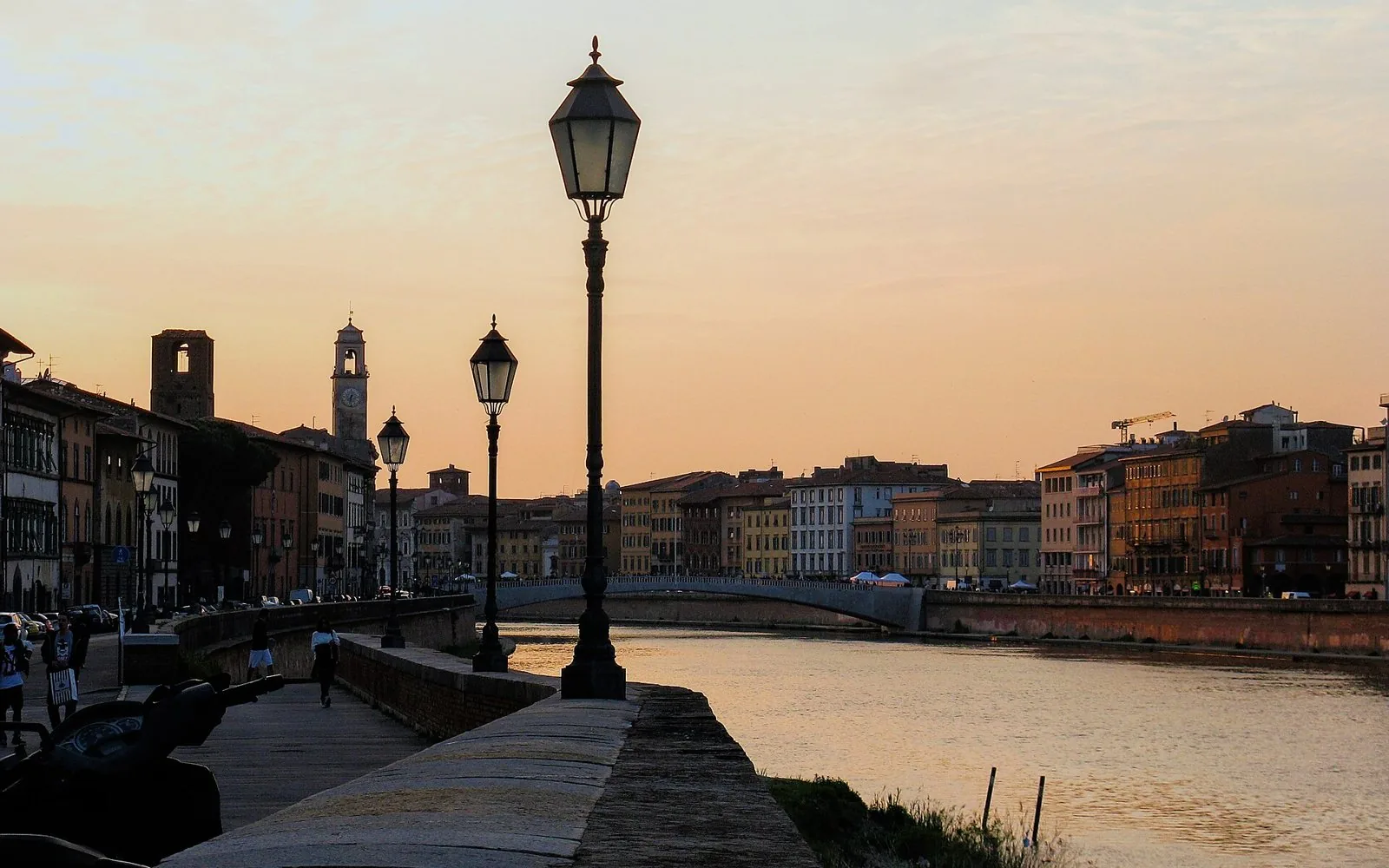 Il Lungarno di Pisa al tramonto con i lampioni, le facciate dei palazzi e un ponte sull'Arno sullo sfondo